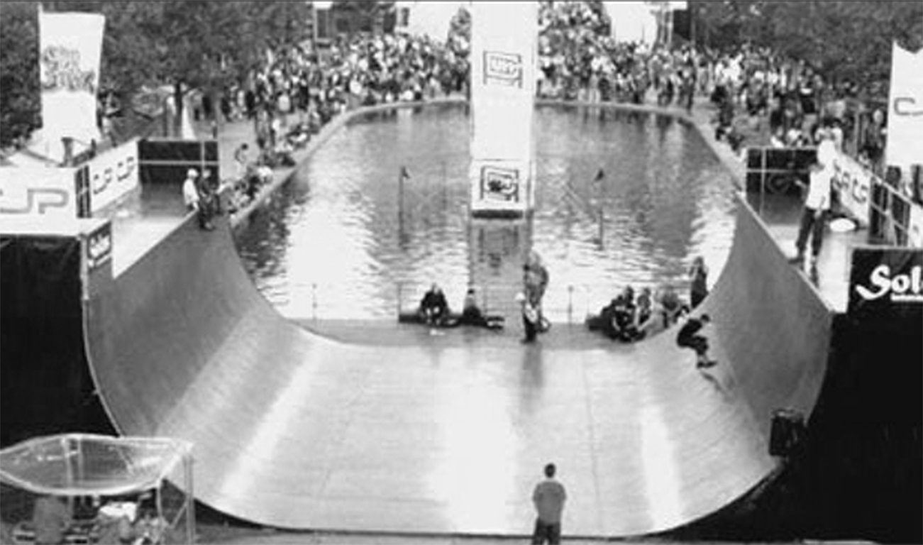 Portable vert ramp set up at an outdoor event, with spectators and a water feature behind